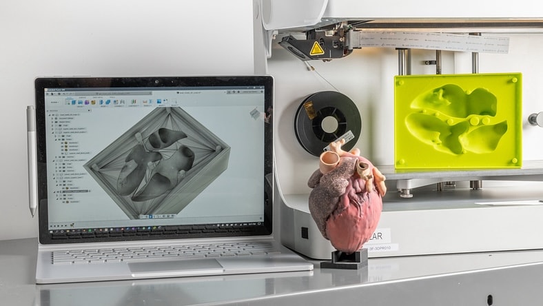 On a table in a lab sits a laptop displaying 3D-modeling software, a model of a human heart, a 3D printer, and a 3D-printed prototype for enclosing a heart for transplant.