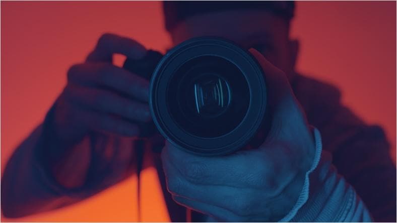 A close-up looking into the lens of a camera being held by a person in front of red backlighting.