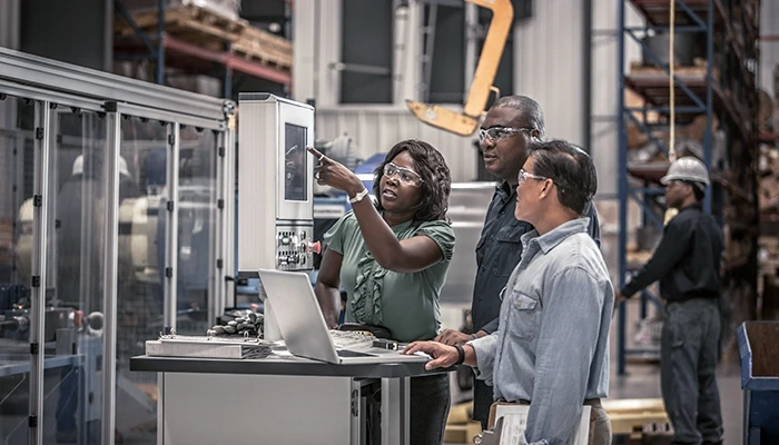 Three people in safety glasses look at a machine in a factory.