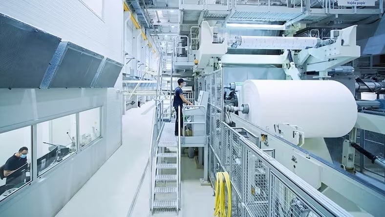 A view of an automated paper milling factory shows enormous rolls of paper on machinery, a worker on an elevated platform monitoring operations, and windows into an office where a facilities manager works on computers.