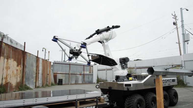 A Cosmic Robotics prototype extends an arm equipped with suction end-effectors toward a stack of solar panels.