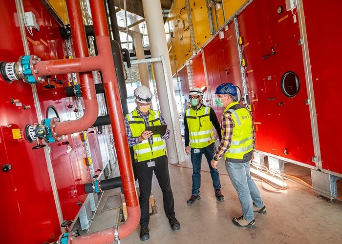 Three men in work vests, hard hats, and masks in a technical room