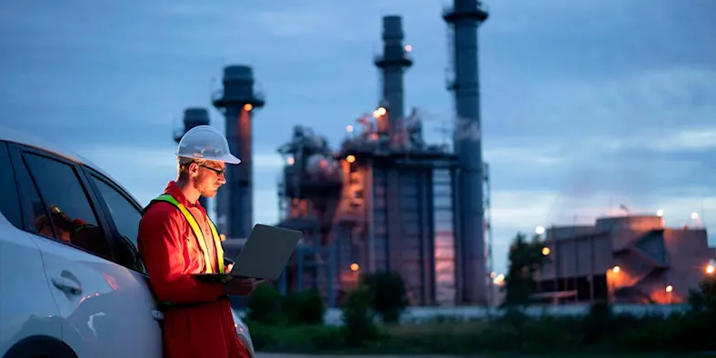 A man in a construction suit and hard hat leans on a car outside of a power plant at dusk.