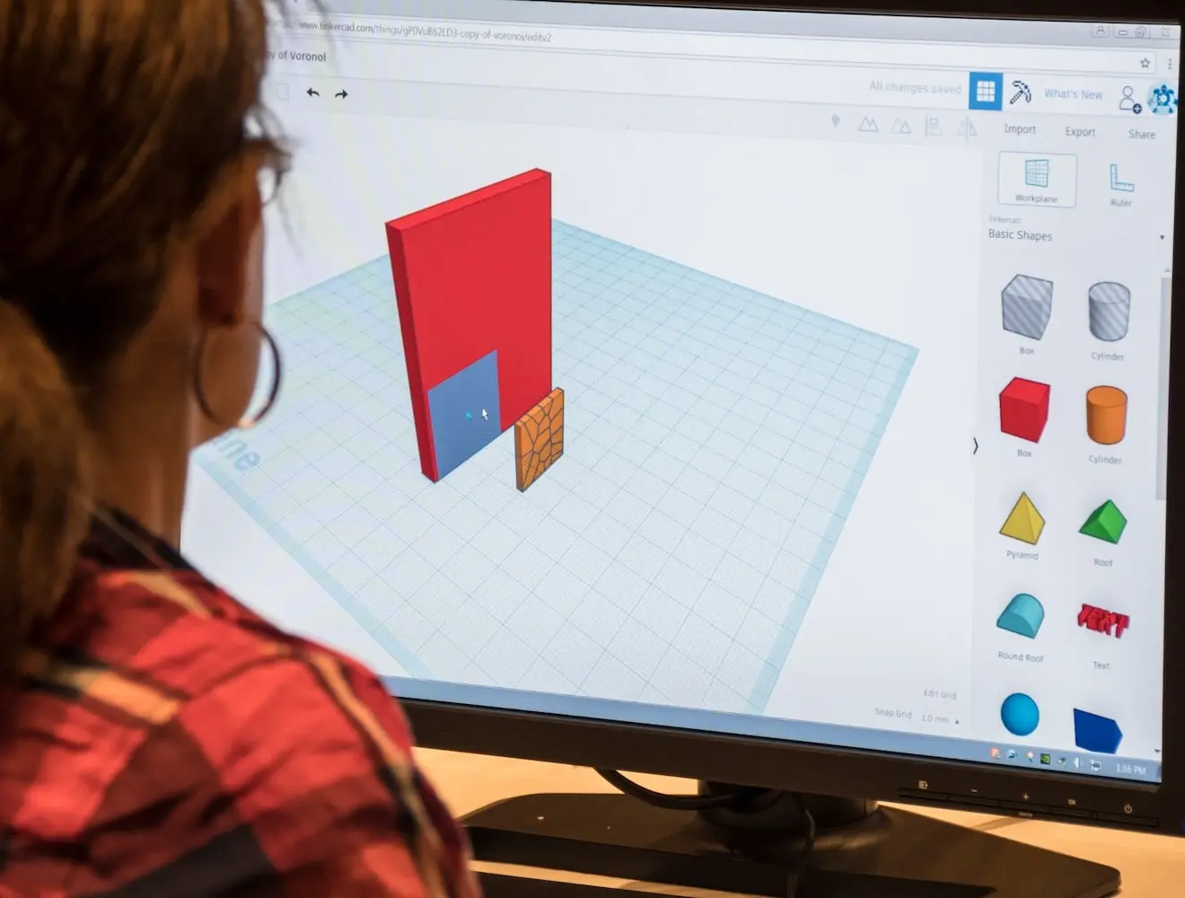 A view over the shoulder of a woman shows her using TinkerCAD on a desktop computer.