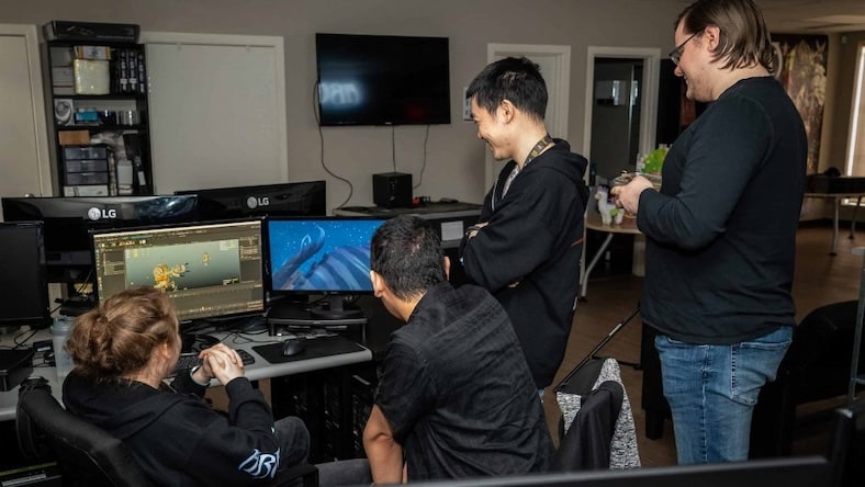 Four people review 3D models on computer screens in an office.