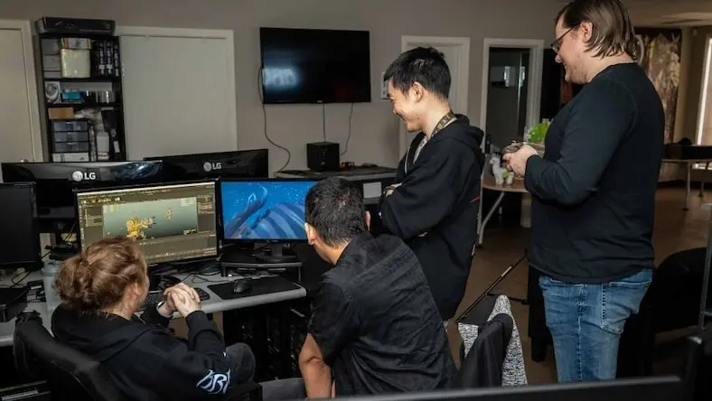 Four people review 3D models on computer screens in an office.