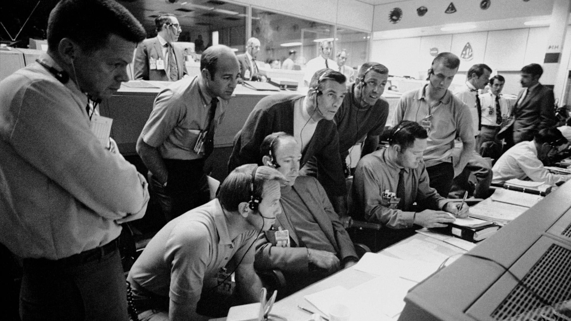 In this black and white photo, Two dozen NASA engineers gather around monitoring equipment during the Apollo 13 moon landing. 
