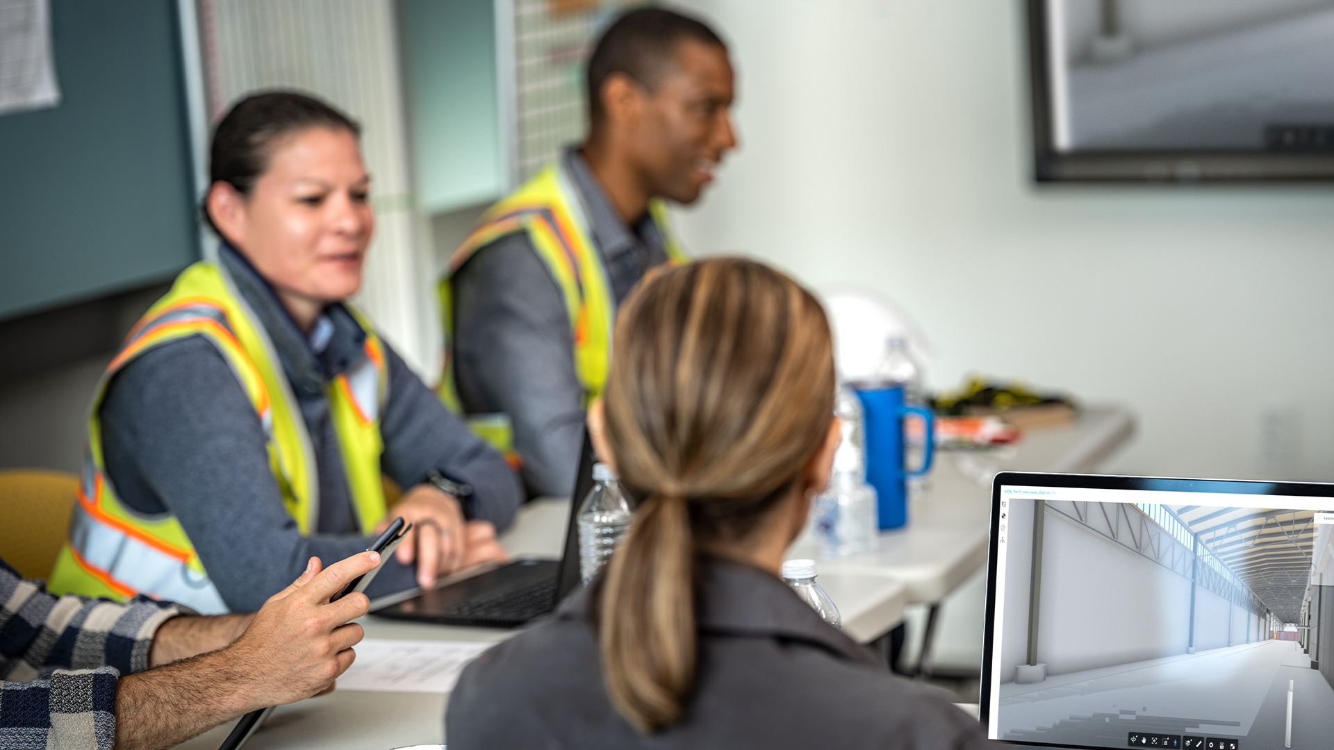 Three people sitting at a table with one person holding a smartphone and another person viewing a laptop screen showing an empty warehouse interior
