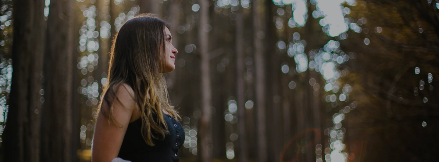 A young woman  with brown hairs standing in woods.