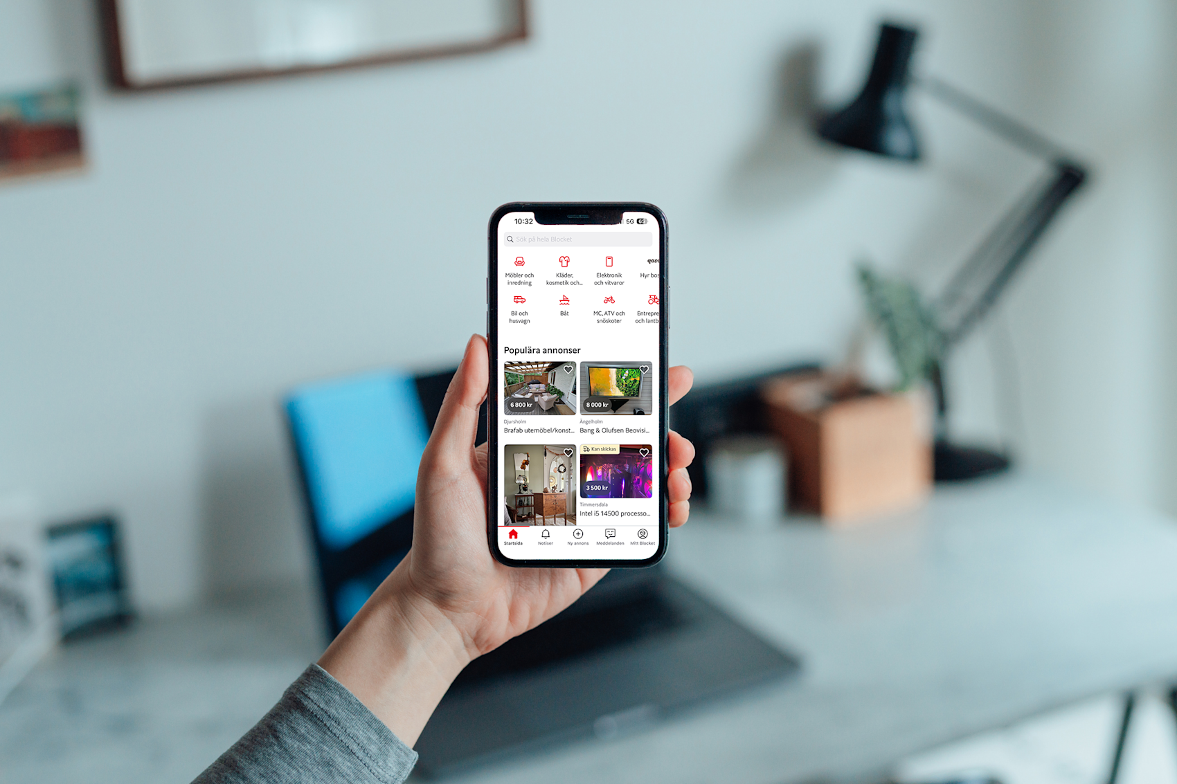 Hand som. håller en mobiltelefon med Blocket-appen öppen, som visar populära annonser och kategorier i en suddig inomhusmiljö.