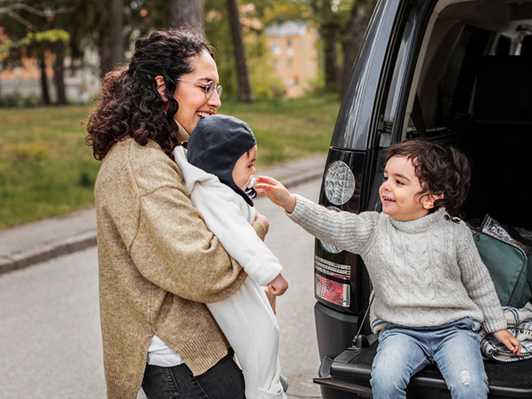 Kvinna står med två barn vid baklucka på bil.