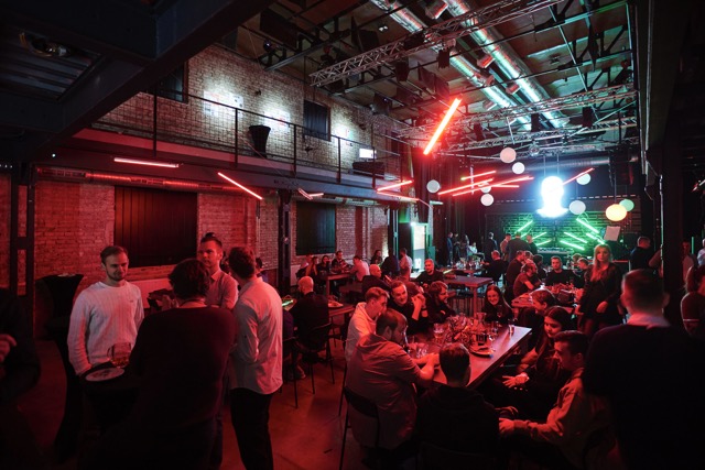 A lively indoor event with people socializing at tables under red neon lights, exposed brick walls, and a brightly lit stage in the background. The atmosphere is energetic and festive.