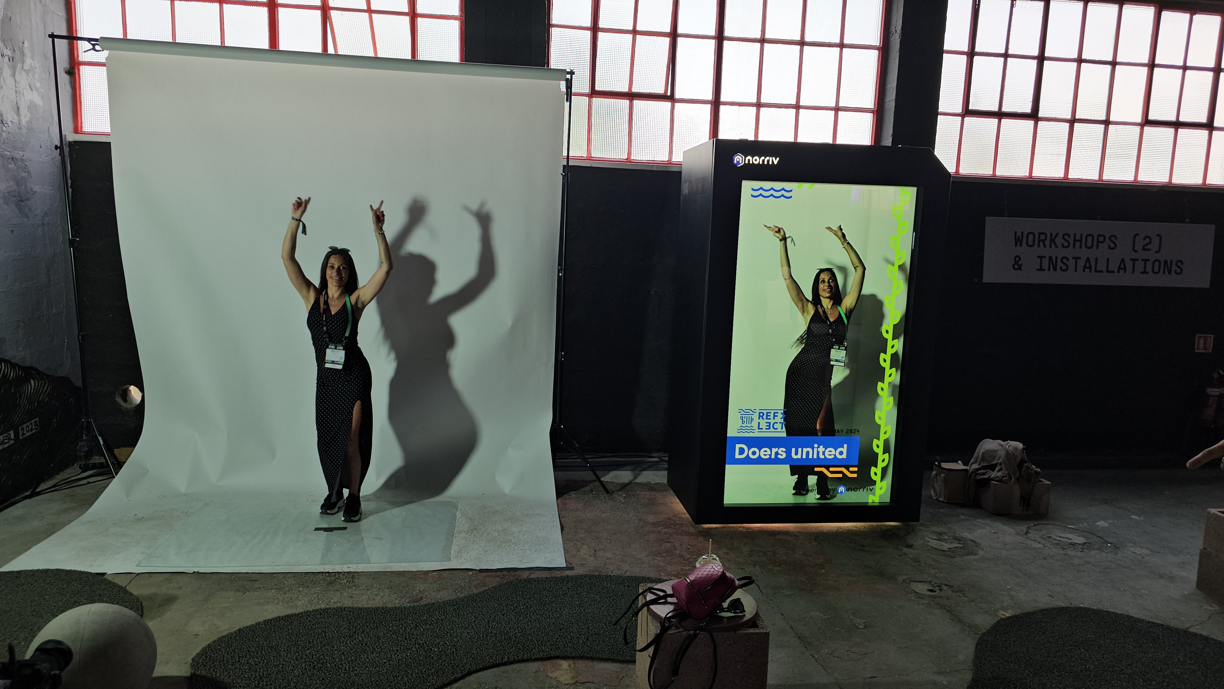 A woman poses with arms raised in front of a white backdrop, casting a shadow. Next to her is a large digital screen displaying her image with the text Doers united. Industrial windows are in the background.