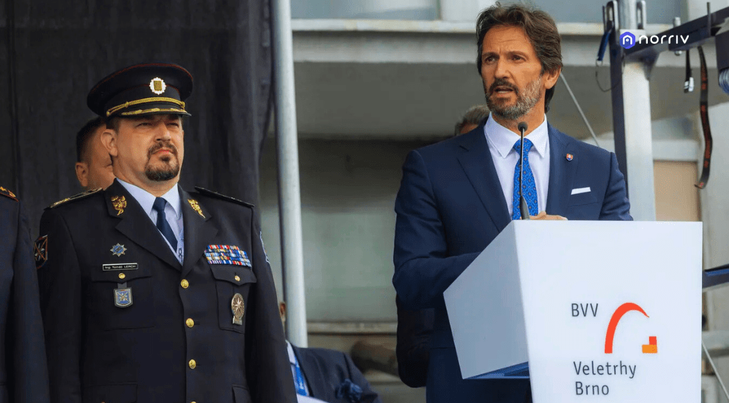 A man in a police uniform stands beside a man in a suit speaking at a podium labeled BVV Veletrhy Brno during an outdoor event.