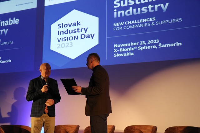 Two men stand on stage under projected screens at Slovak Industry Vision Day 2023. One holds a microphone, the other a folder. Event details and date, November 23, 2023, are visible on the screen behind them.