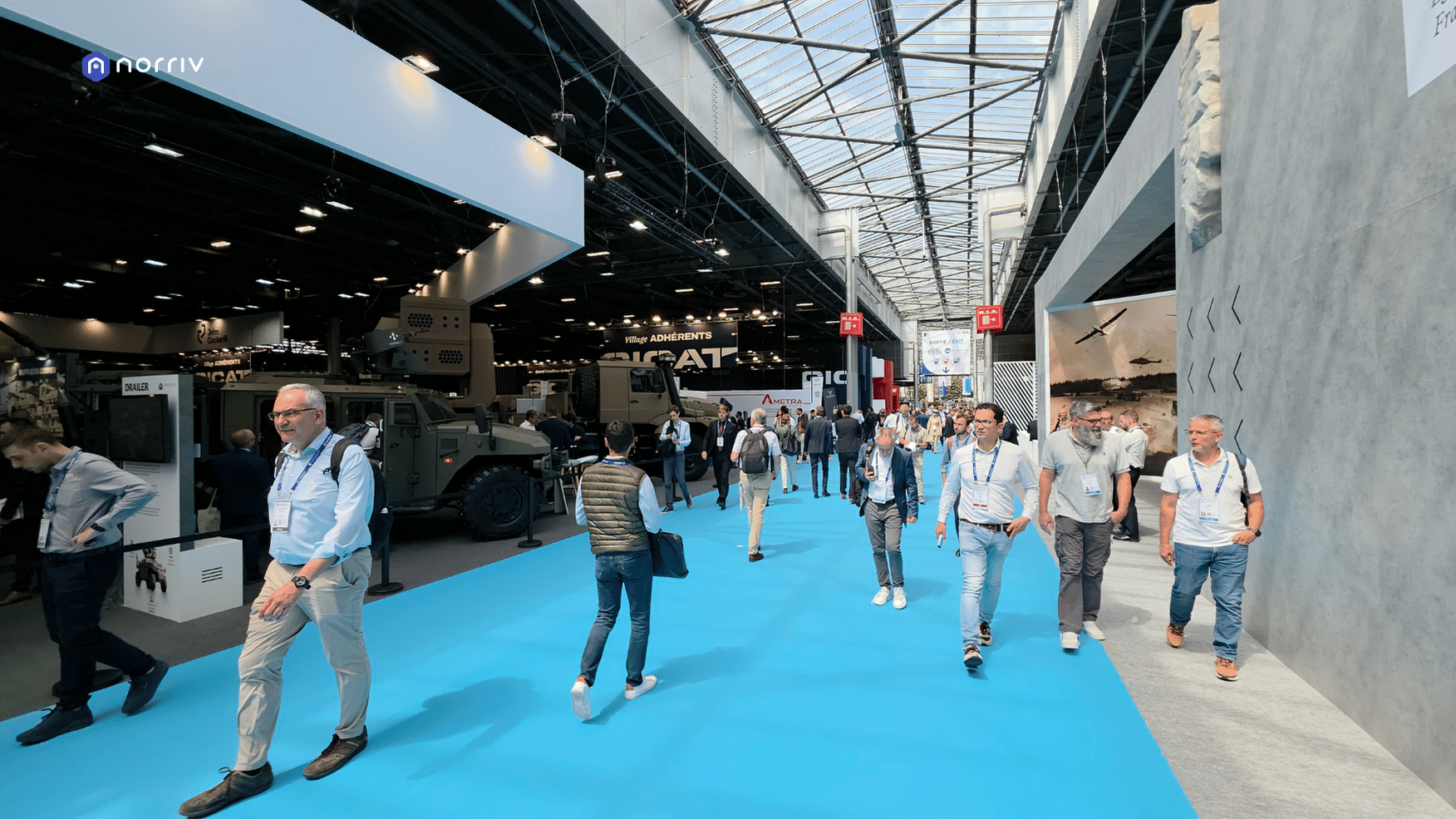 Visitors walking along a blue-carpeted aisle between defense exhibition booths and military vehicle displays at Eurosatory 2026.