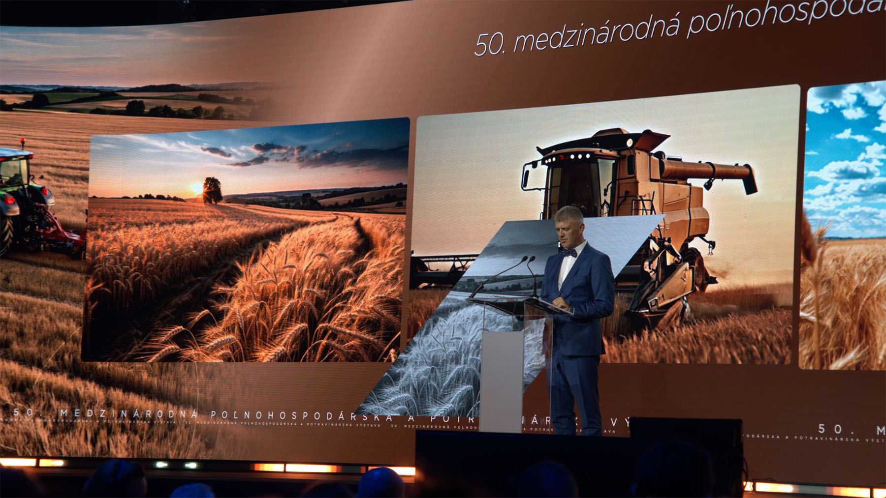 A man in a suit speaks at a podium on stage, with wheat fields and combines shown on a large screen behind him.