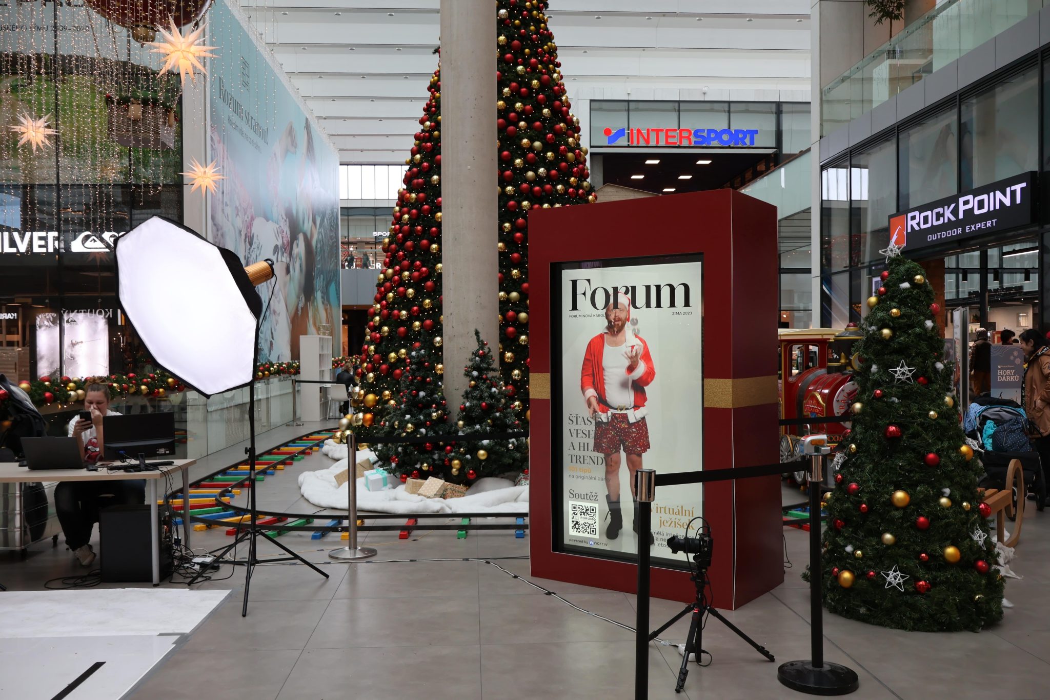 A shopping mall decorated for Christmas with trees, ornaments, and a large norriv Hologram Box display, surrounded by lighting equipment.