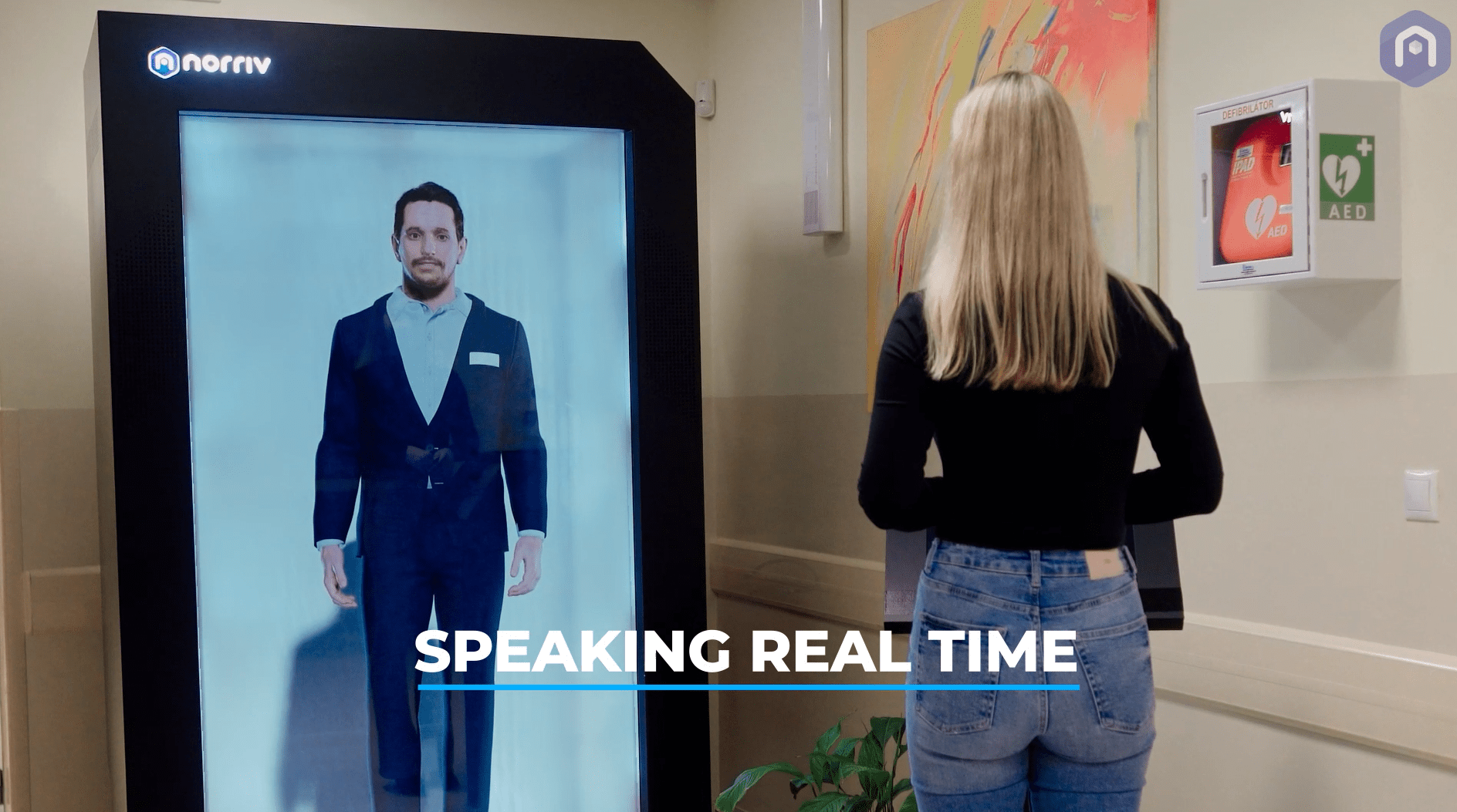A woman stands in front of a large digital screen displaying a life-sized man in a suit, with the words SPEAKING REAL TIME written across the bottom. The setting appears to be indoors, possibly in a public building.