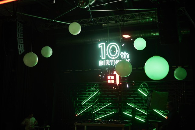 A dark room decorated with white balloons and bright green neon lights displays a sign reading “10th Birthday.” The atmosphere suggests a festive celebration or party.