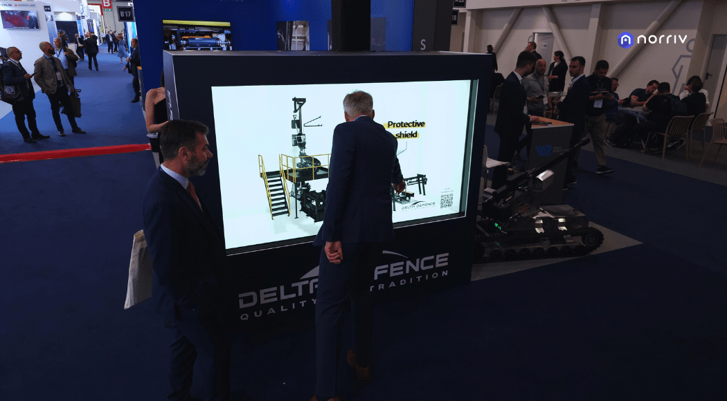 Two men in suits look at a large digital display showing a diagram of protective equipment. People are seated and walking in the background at a convention or exhibition. The setting appears to be a trade show booth.