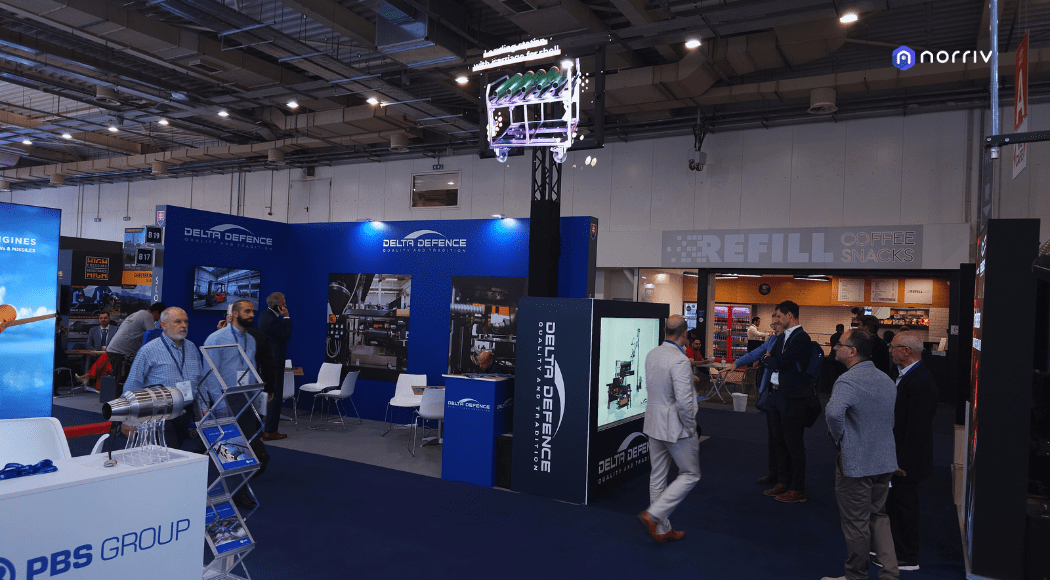 A trade show floor with people talking and walking among various company booths. Blue panels display DELTA DEFENCE branding, and a REFILL Coffee Snacks sign is visible on a wall in the background.