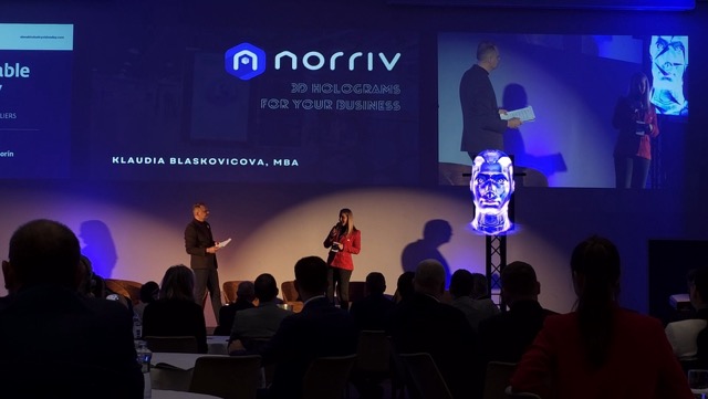 A man and a woman speak on stage at a business event with an audience watching. Behind them is a large screen displaying norriv 3D holograms for your business and Klaudia Blaskovicova, MBA.