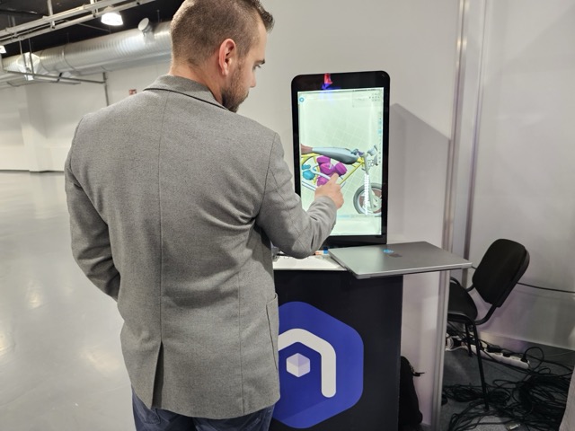 A man in a gray blazer interacts with a large touchscreen displaying a colorful 3D model, standing at a booth in a modern, spacious indoor setting.