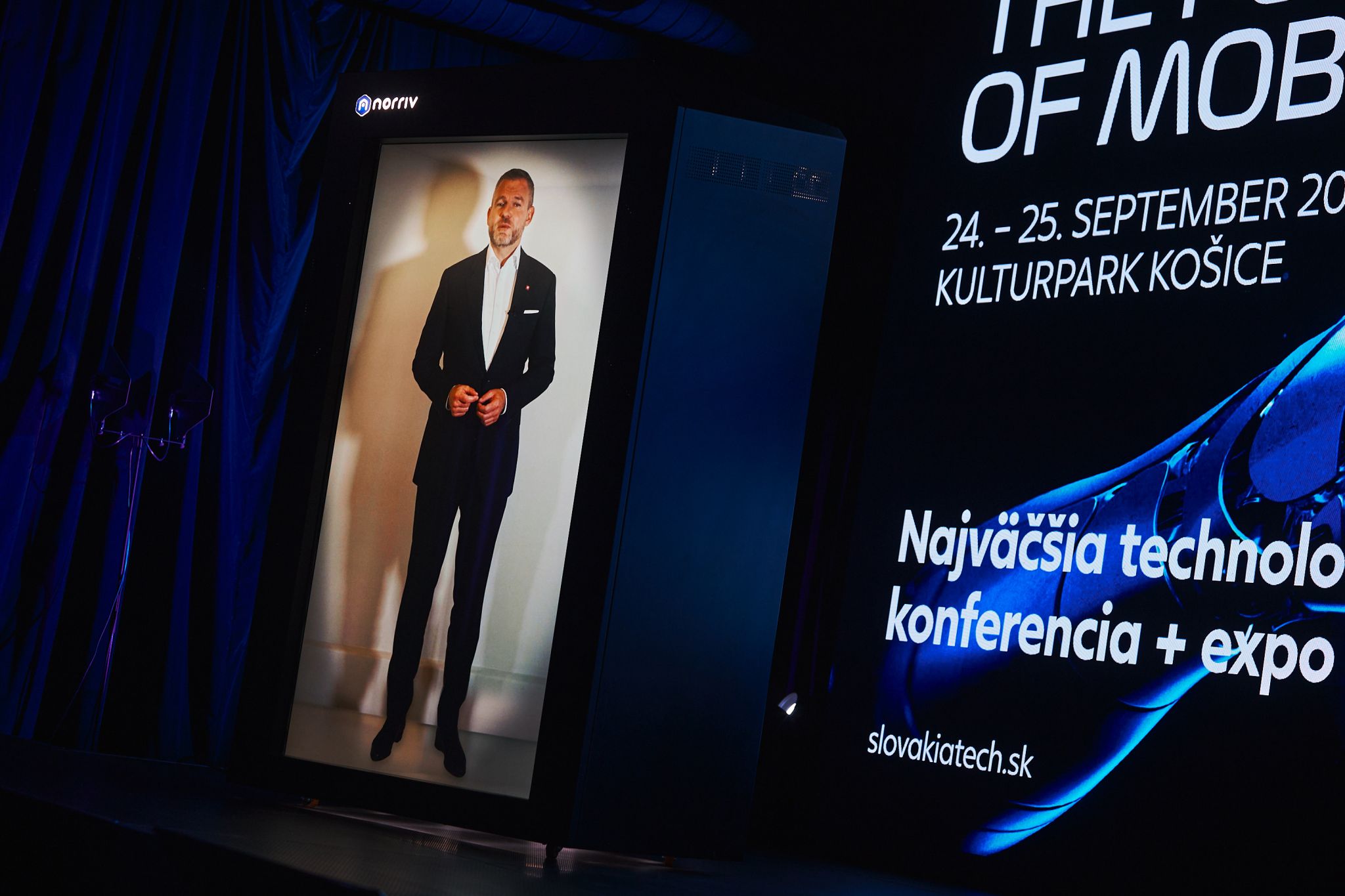 A man in a suit appears as a life-sized hologram on stage at a technology conference, with event details projected on a screen beside him.
