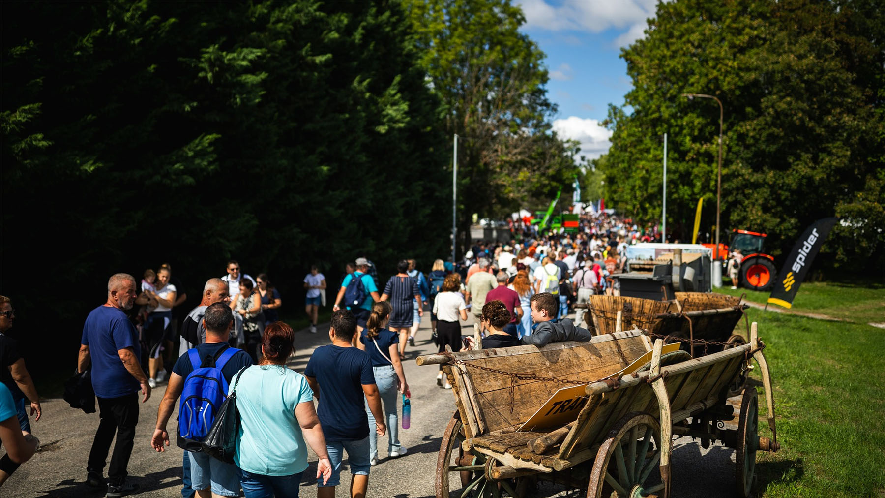 A crowd of people walks down a tree-lined paved road, passing wooden carts and a red tractor beneath a partly cloudy sky.