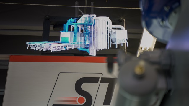 A holographic projection of a complex machine or industrial equipment appears above an ST-branded sign, with part of another mechanical device visible in the foreground on the right.