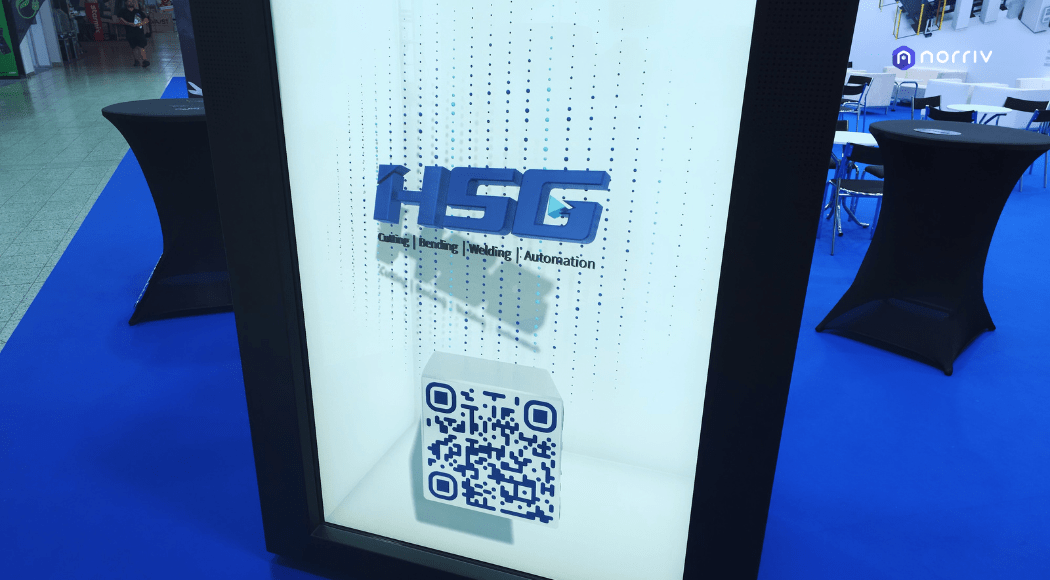 A display panel with the HSG logo and the words Cutting | Bending | Marking | Automation above a QR code, set against a blue carpeted floor and tall black tables in an exhibition booth.