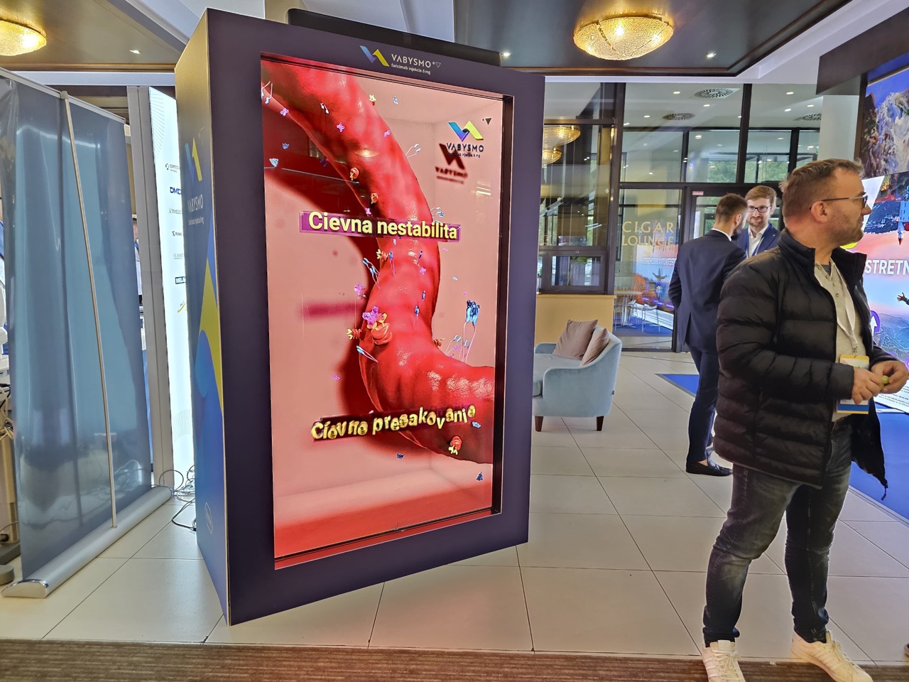 A man stands next to a large interactive display showing a 3D model of a blood vessel with Slovak text. Several people and banners are visible in the background of the modern indoor setting.