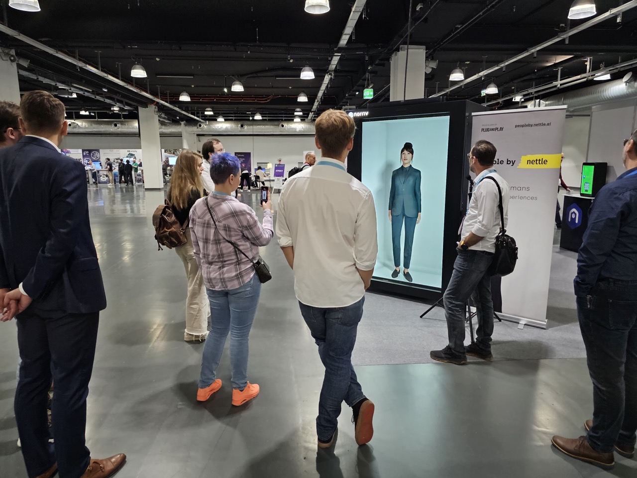 A group of people stands in a modern exhibition hall, watching a large digital screen displaying a full-body image of a person. Exhibition booths and banners are visible in the background.