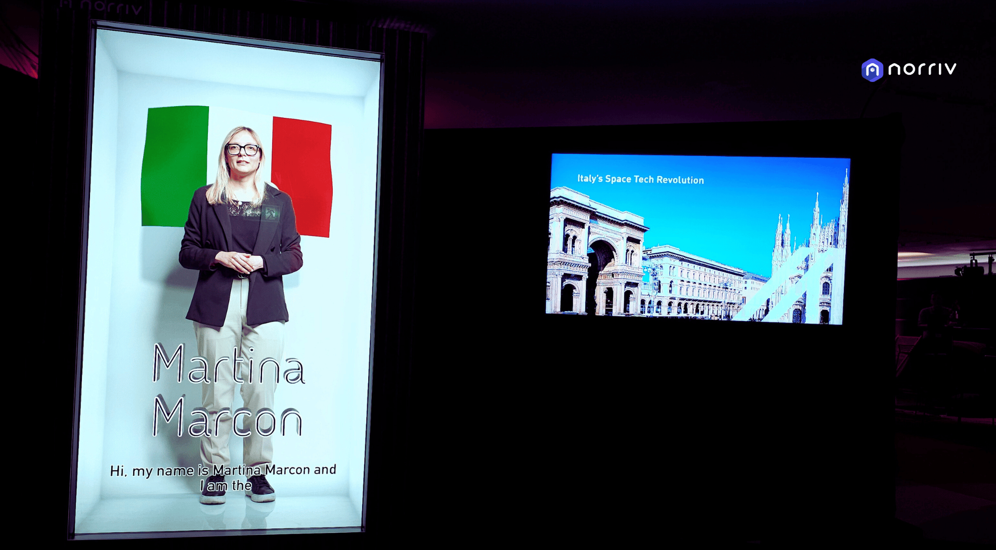 A digital display shows a woman named Martina Marcon standing in front of an Italian flag, with text about her. Beside it, another screen displays Italy’s Space Tech Revolution and an image of Italian architecture.