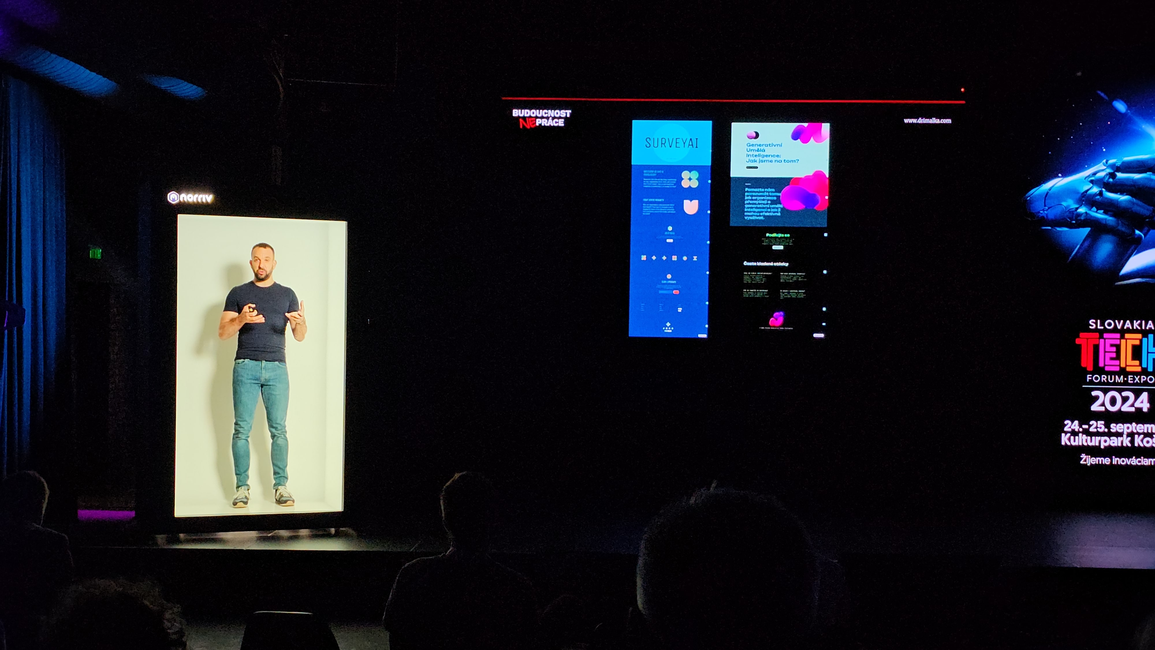 A man appears as a hologram on stage next to large presentation screens displaying graphs, text, and event details at a technology forum in a dark auditorium.