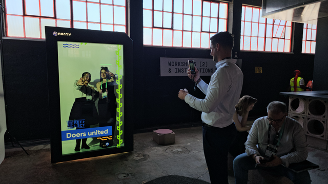 A man interacts with a large digital screen displaying two people posing and the text Doers united. Nearby, other people are seated, with industrial-style windows and a WORKSHOPS & INSTALLATIONS sign in the background.