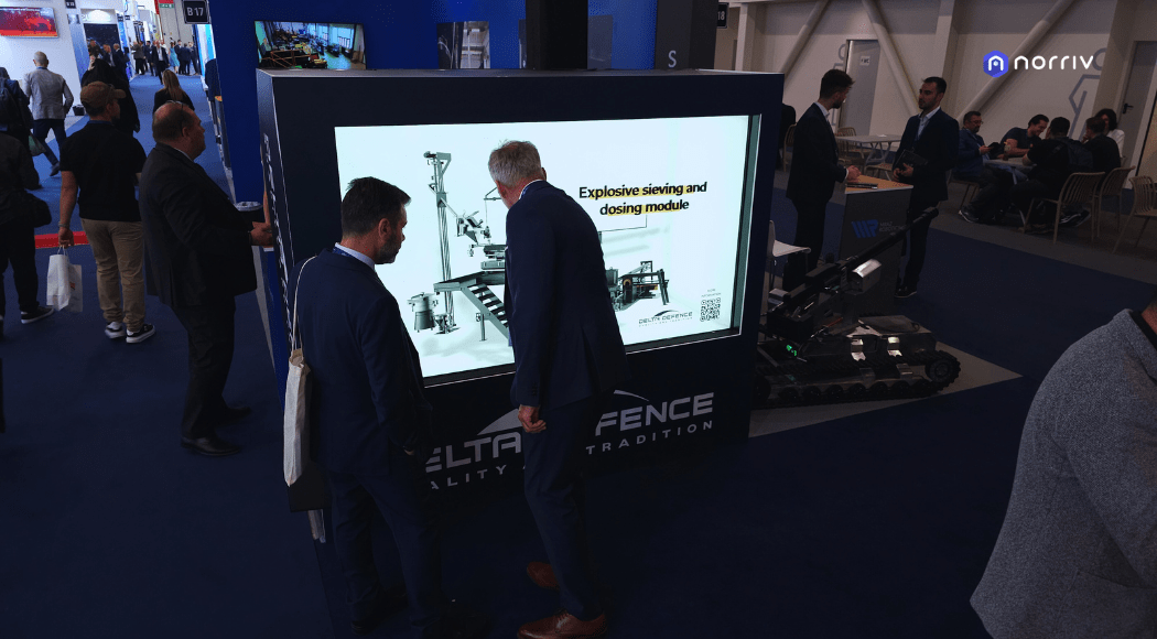 Two men in suits look at a large touchscreen display showing an Explosive sewing and dosing module diagram at a technology trade show with other people and booths in the background.