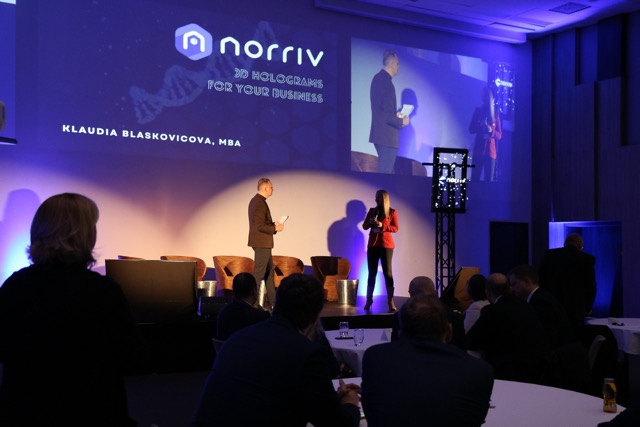 Two people stand on stage under spotlights, with a projected logo reading norriv 3D Holograms For Your Business and Klaudia Blaskovicova, MBA behind them. Audience members sit at round tables in the foreground.