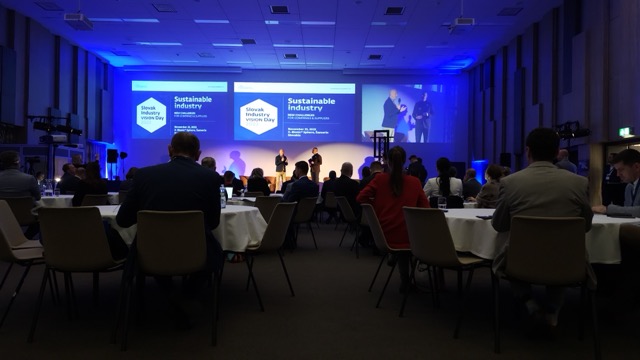 People seated at round tables in a dimly lit conference room, watching a presentation on large screens labeled “Sustainable Industry.” Blue lighting highlights the stage where presenters are speaking.