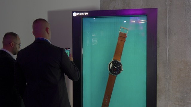 Two men in suits stand in front of a large digital display showing an image of a wristwatch with a brown leather strap. One man appears to be taking a photo of the display with his phone.