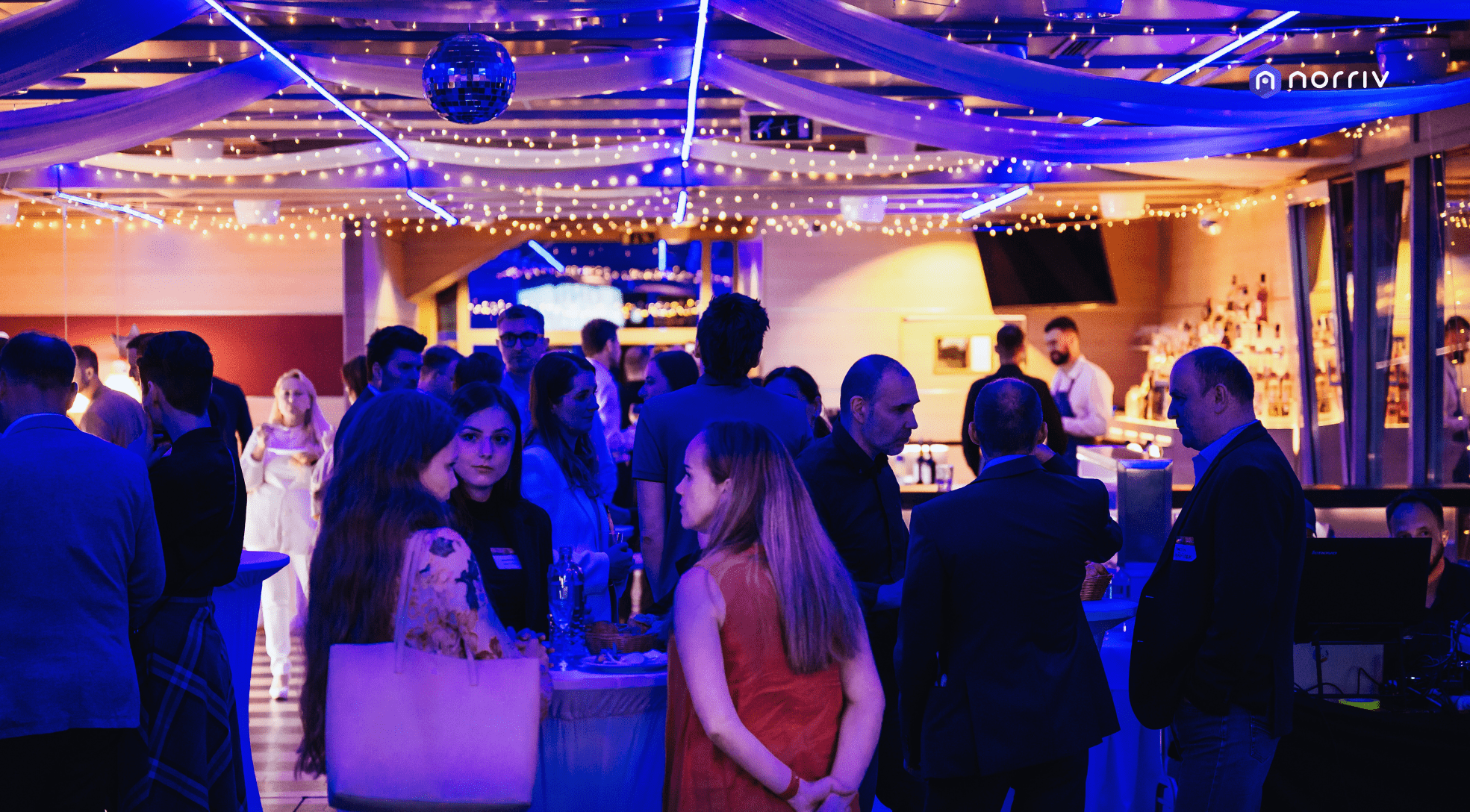 A lively indoor event with people socializing under strings of blue and white lights and fabric drapery. Guests stand around cocktail tables near a bar, creating a festive, elegant atmosphere.