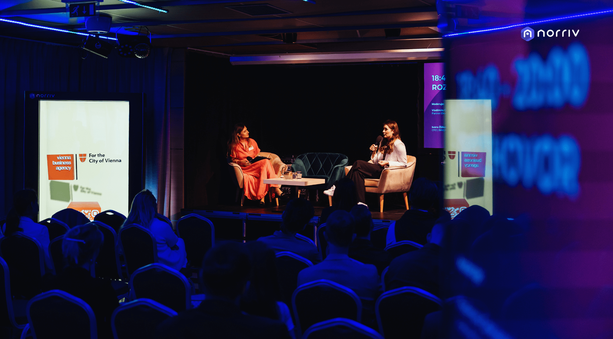 Two women sit on stage in armchairs having a discussion at a conference, with an audience seated in front of them. Large screens display Vienna Business Agency and For the City of Vienna logos.