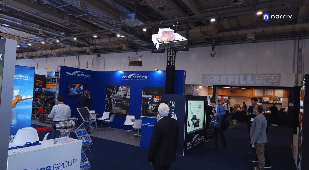 People attend a trade show with booths featuring technology and aerospace displays. Several men in business attire walk around, while a REFILL Coffee Snacks stand is visible in the background.