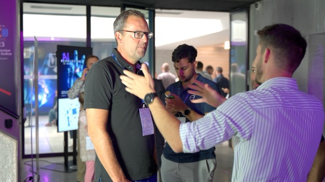 A group of men are interacting at an indoor event. One man in front gestures while talking to another wearing a black shirt and glasses. Other people are visible in the background, some using their phones.