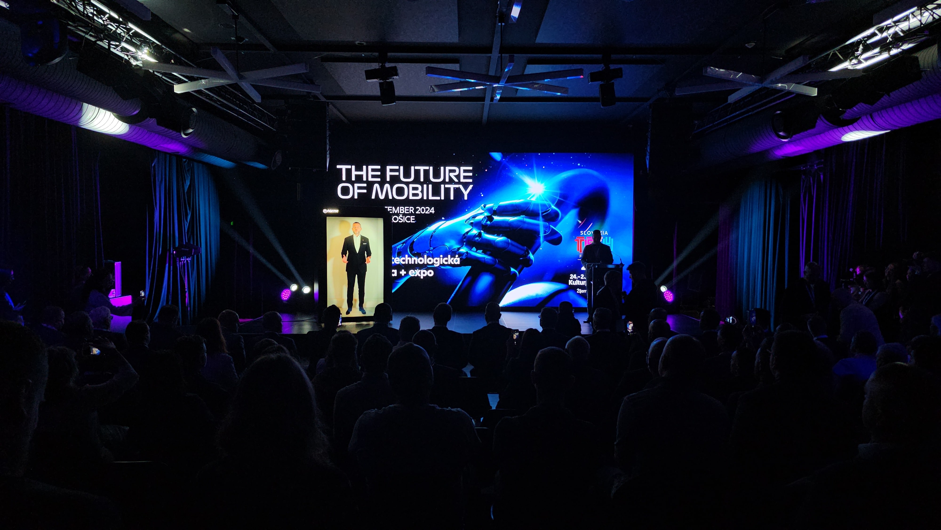 A dark conference room filled with people facing a brightly lit stage. Onstage, a person stands next to a large digital screen displaying the words “The Future of Mobility” and an image of a robotic hand touching a human hand.