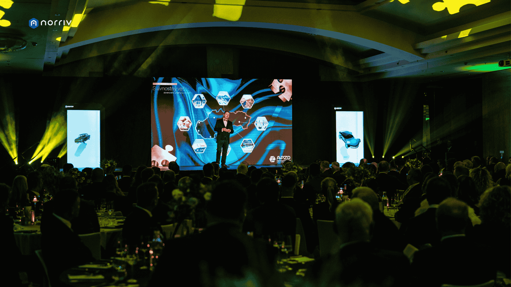 A speaker stands on stage in front of a large, colorful screen at a conference or gala event, with an audience seated at round tables in a dimly lit, elegant venue.