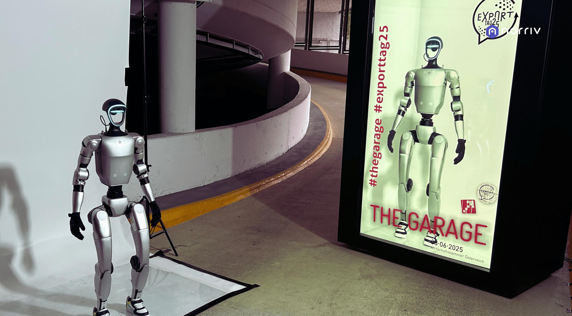 A humanoid robot stands on a mat in a parking garage next to a large advertisement display showing the same robot and the words “THE GARAGE” with hashtags and event details.