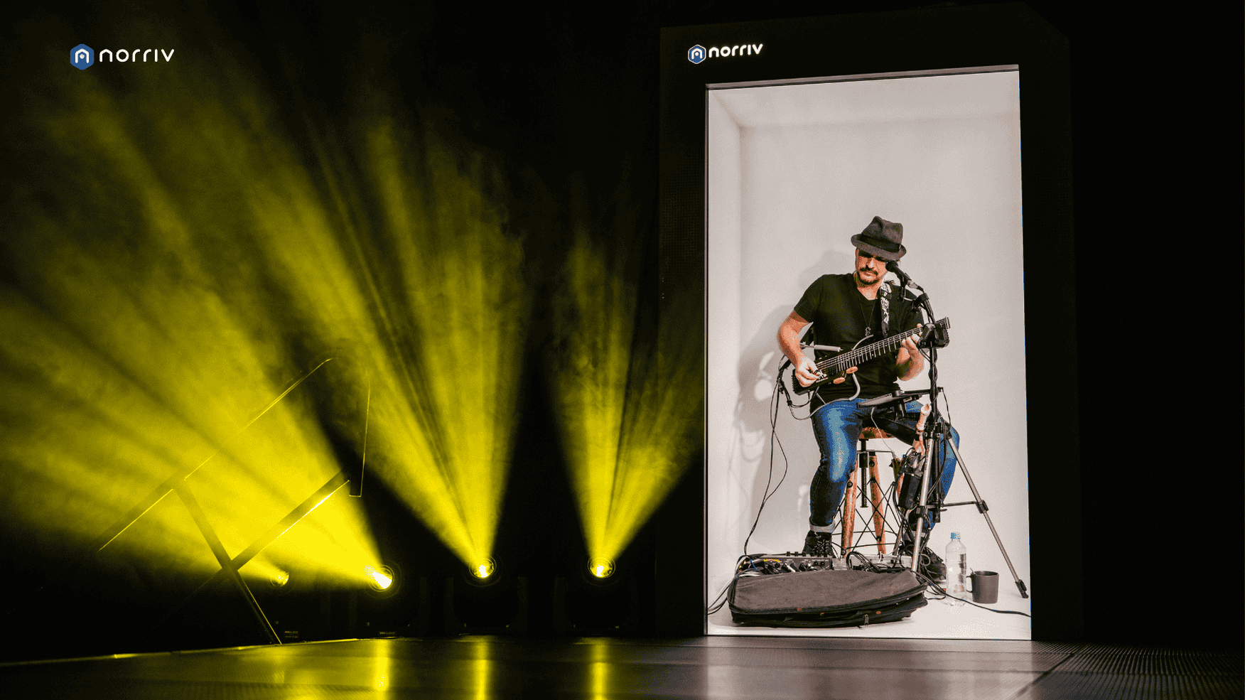 A musician wearing a hat and sunglasses plays an electric guitar while seated inside a large, vertical, illuminated display. Yellow stage lights shine across the dark stage next to the display.