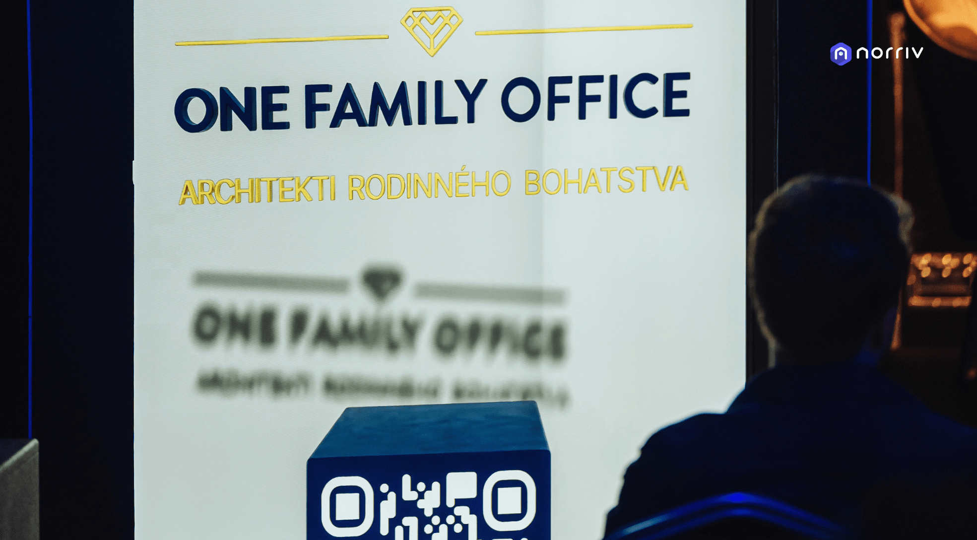 A person sits facing a brightly lit sign that reads ONE FAMILY OFFICE and ARCHITEKTI RODINNÉHO BOHATSTVA, with a QR code visible in the foreground.